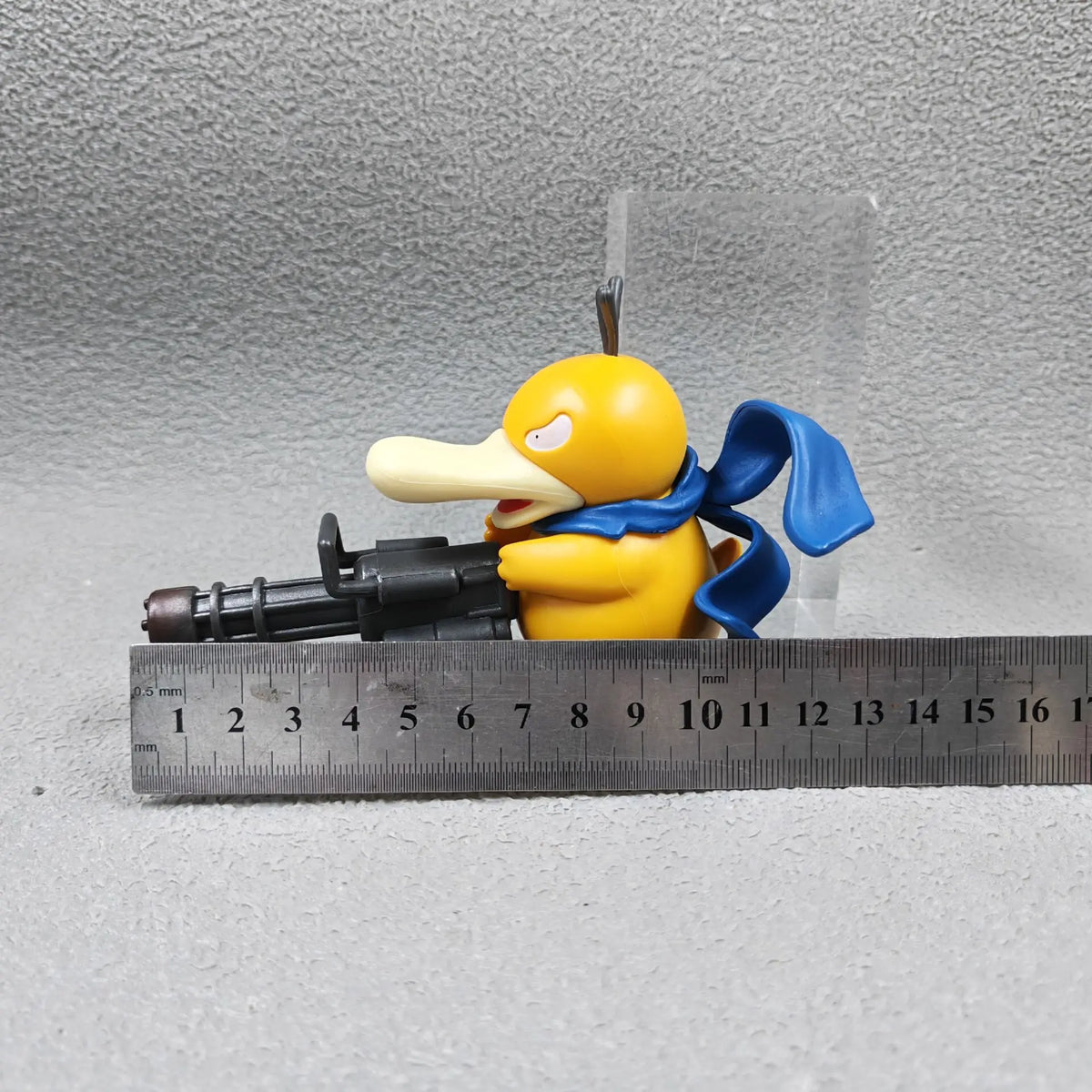 A yellow duck figurine with a blue scarf holds a black Gatling gun, with a metal ruler placed below it.