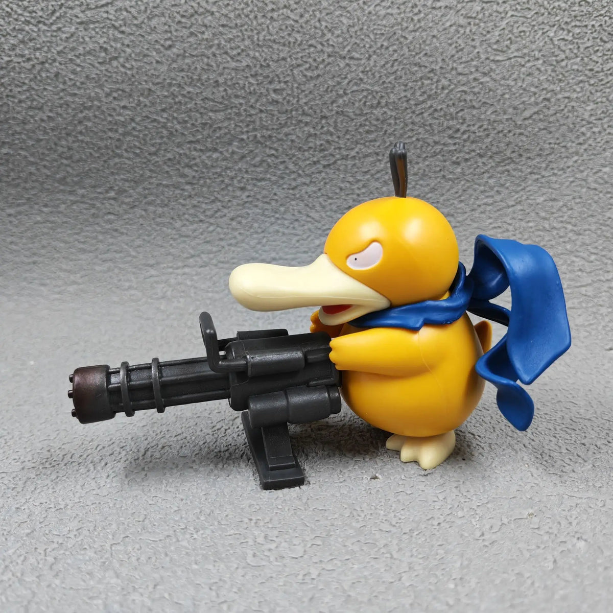 Yellow duck-like figurine with a blue scarf, holding a large black Gatling gun.