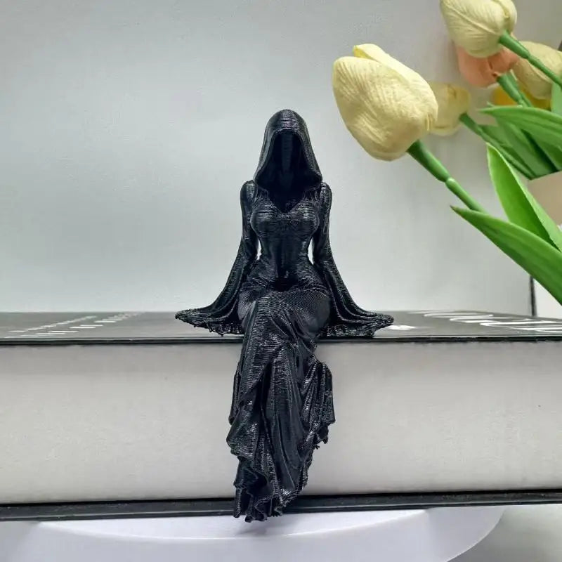 Black seated female grim reaper sculpture with hooded cloak, perched on a book ledge beside yellow tulips.