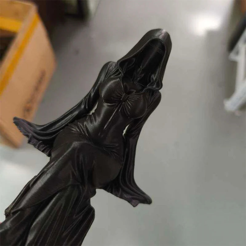 Black figurine of a hooded, robed female figure sitting in a flowing dress with flared sleeves.