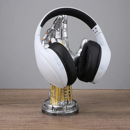 A silver and gold robotic hand stand holds white and black over-ear headphones on a wooden surface.