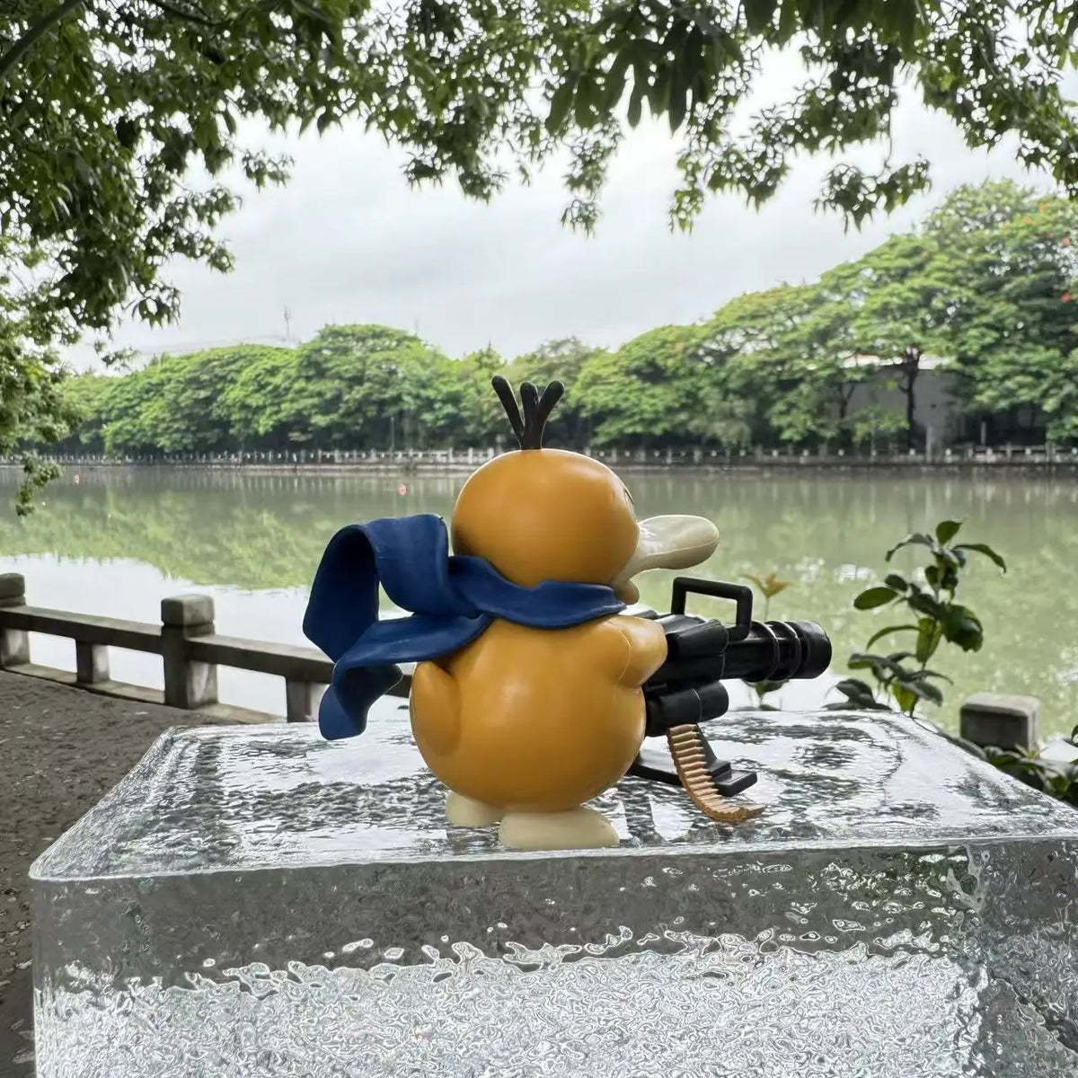 Yellow duck figurine with a blue scarf holding a black Gatling gun, outdoors.