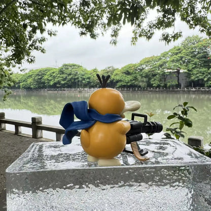 Yellow duck figurine with a blue scarf holding a black Gatling gun, outdoors.
