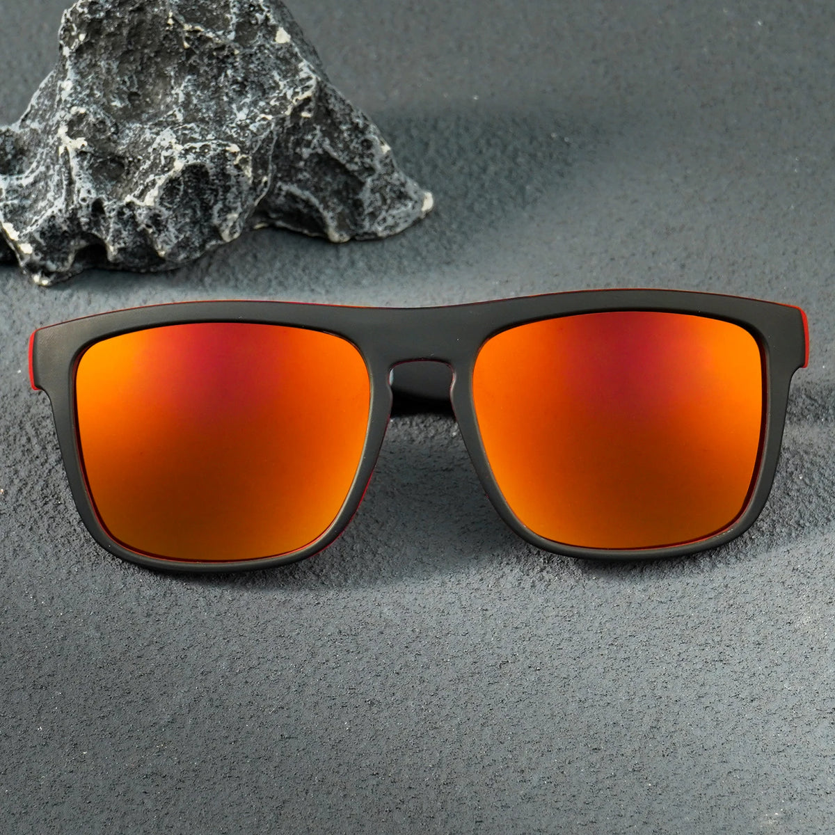 Black square sunglasses with orange-red reflective lenses resting on a dark gray textured surface next to a gray rock.