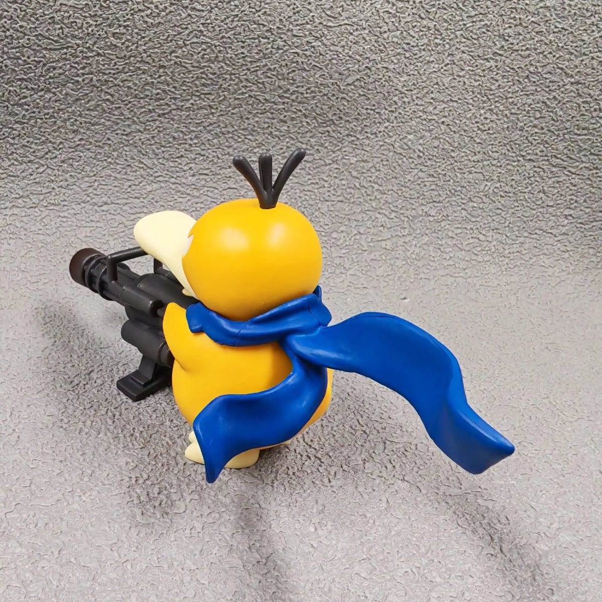 Yellow duck figurine wearing a blue scarf and holding a black Gatling gun.