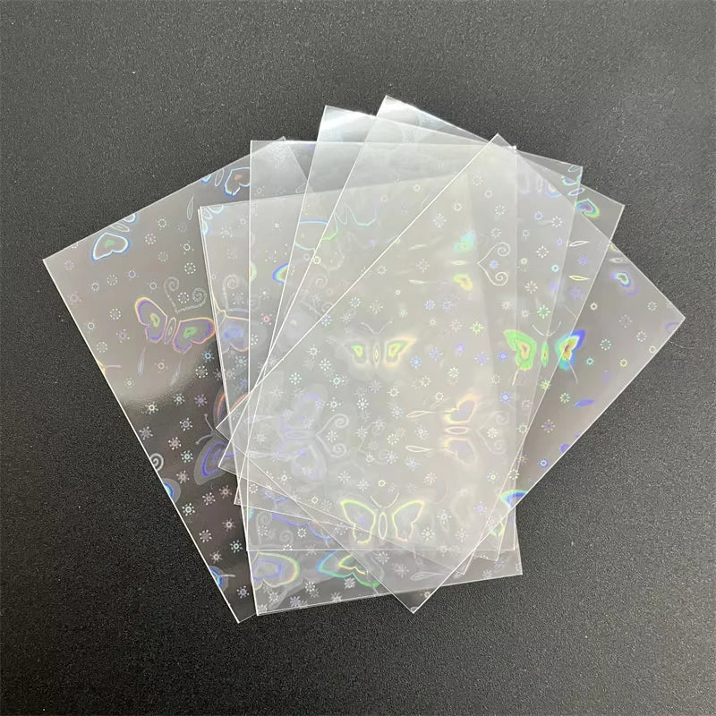 Clear holographic card sleeves featuring a laser butterfly and floral pattern, stacked on a dark surface.