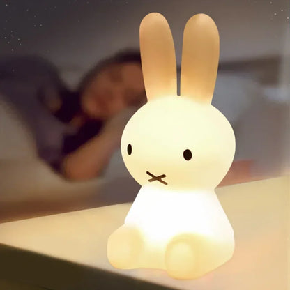Miffy rabbit night light, white and glowing, sits on a surface with a blurred person sleeping in the background.
