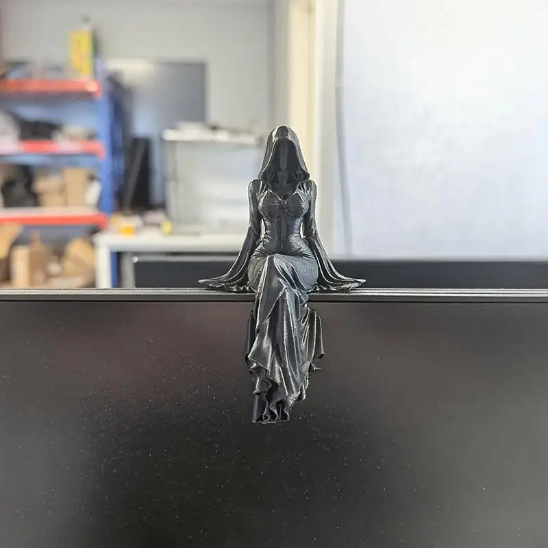 A black resin statue of a hooded, robed female figure sitting with legs crossed on a surface edge.