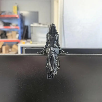 A black resin statue of a hooded, robed female figure sitting with legs crossed on a surface edge.
