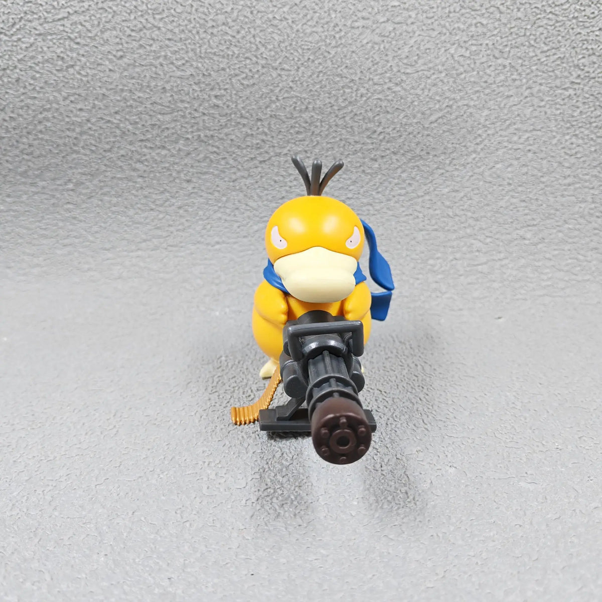 A yellow Psyduck figurine holds a large grey gatling gun with an ammo belt, wearing a blue scarf.