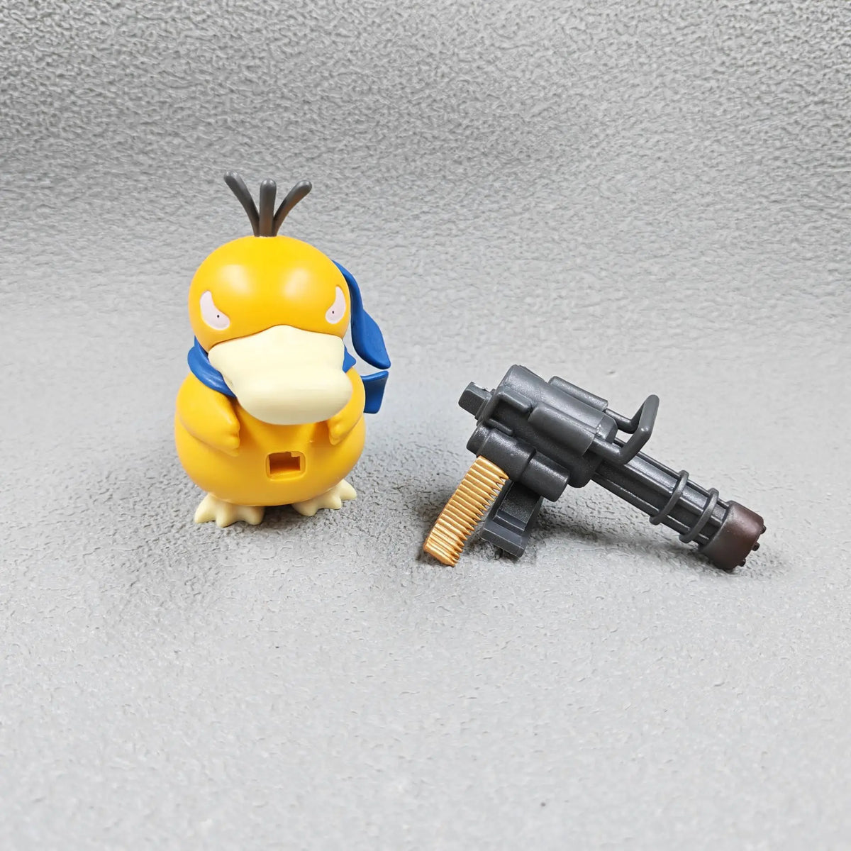 Yellow Psyduck figurine wearing a blue scarf and a gray miniature Gatling gun accessory.