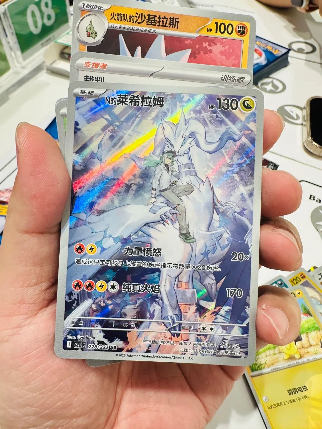 A hand holds a shiny Chinese Pokemon card of N riding Reshiram with glowing blue and white energy.