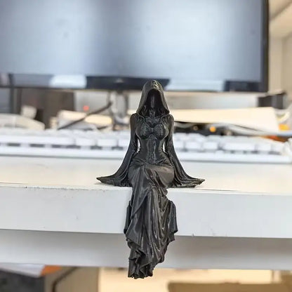 A black, hooded female grim reaper figurine sitting on a ledge with legs dangling.