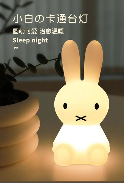 A glowing Miffy rabbit night light with black dot eyes and an X-shaped mouth, sitting beside a potted plant.