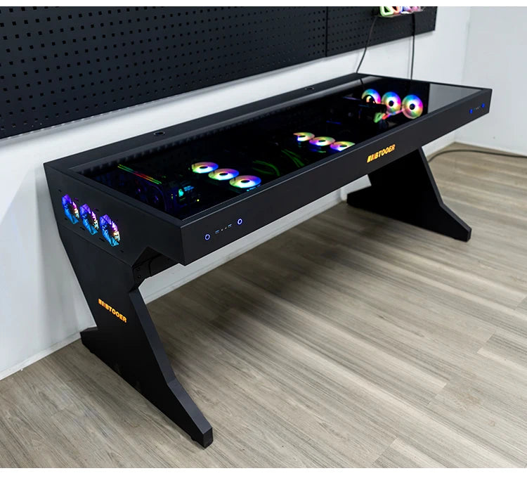 A large black gaming desk with an integrated glass-top PC case showcasing vibrant RGB fans and hardware.