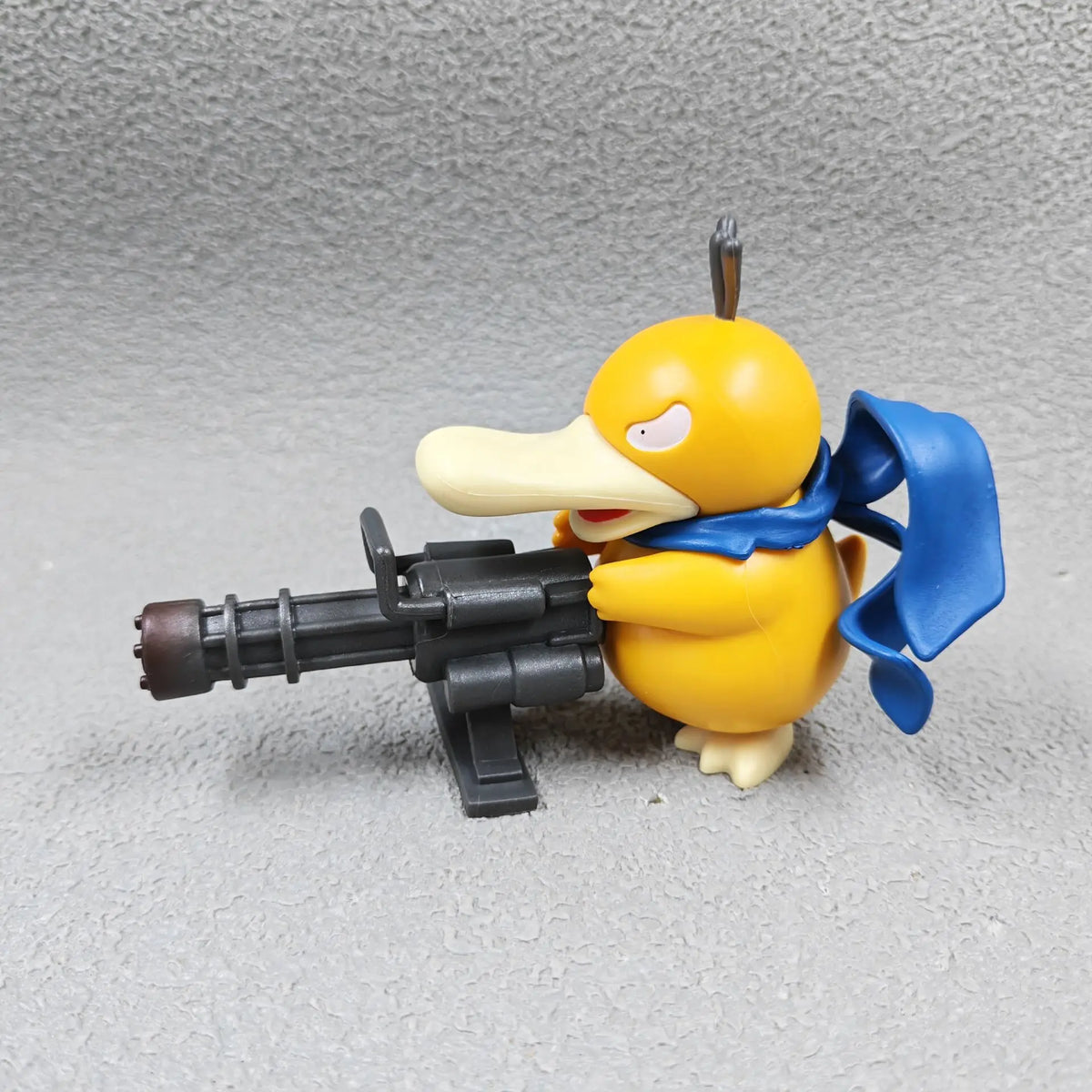Yellow duck-like figurine with a blue scarf, wearing a stern expression and holding a large gray Gatling gun.