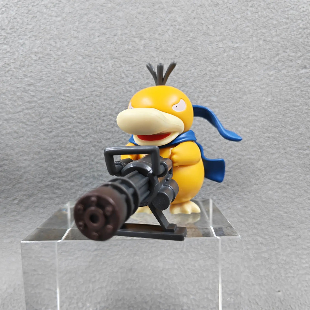 An orange duck-like figure with a blue scarf holds a black Gatling gun, standing on a clear block.