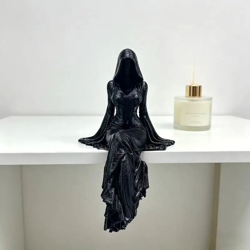 A black, hooded female figure statue sits on a white surface with legs dangling.