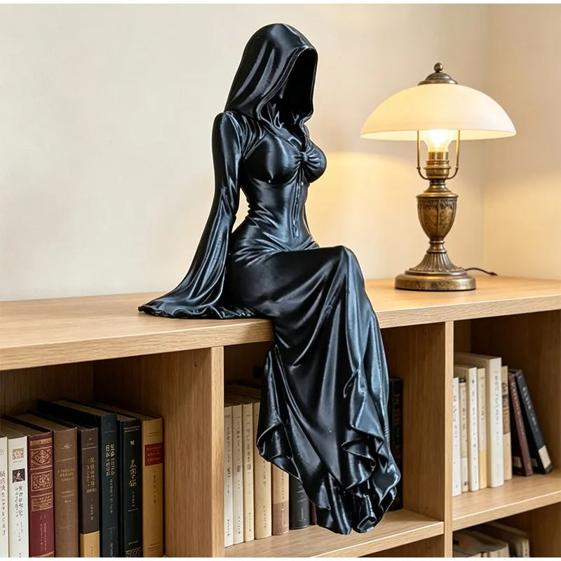 Black-hooded, sitting female statue with flowing robes, posed on a wooden shelf next to a lit lamp.