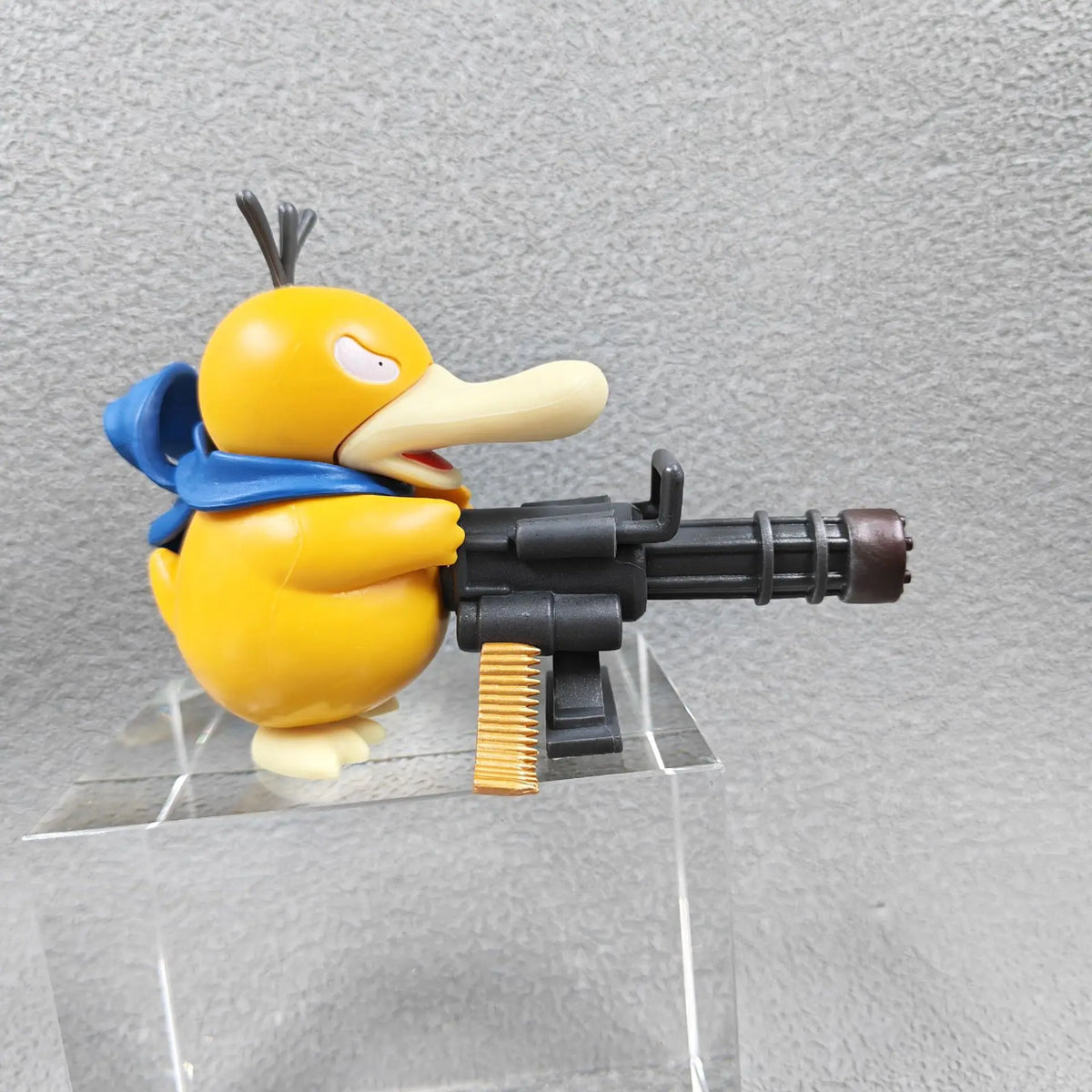 Yellow Psyduck figurine wearing a blue scarf and holding a black Gatling gun with gold bullets on a clear stand.