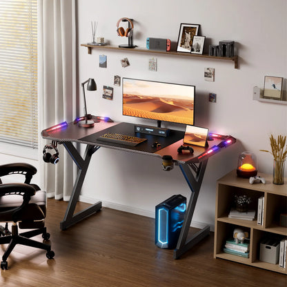 Black Y-shaped gaming desk with RGB LED lights on the surface, supporting a monitor, keyboard, and mouse.