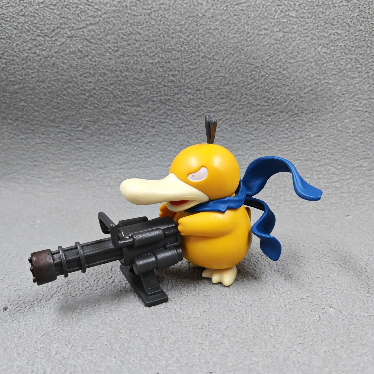 Yellow duck-like figurine with a blue scarf, holding a large black Gatling gun.