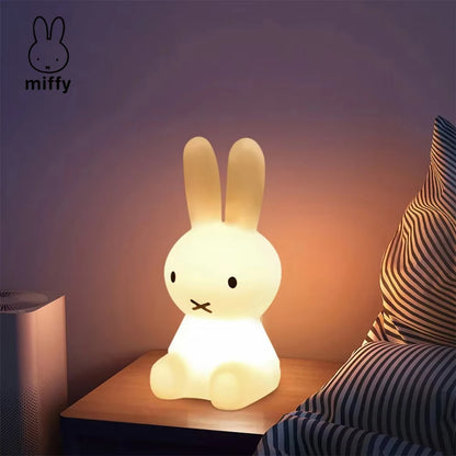 Miffy rabbit night light glowing softly on a bedside table next to striped pillows.
