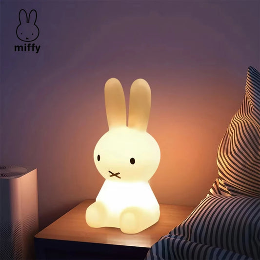 Miffy rabbit night light glowing softly on a bedside table next to striped pillows.