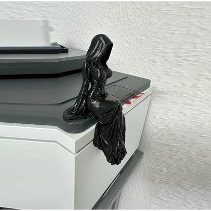 A black, hooded female reaper figurine sits on the edge of a white and gray desk, with her gown draped down.
