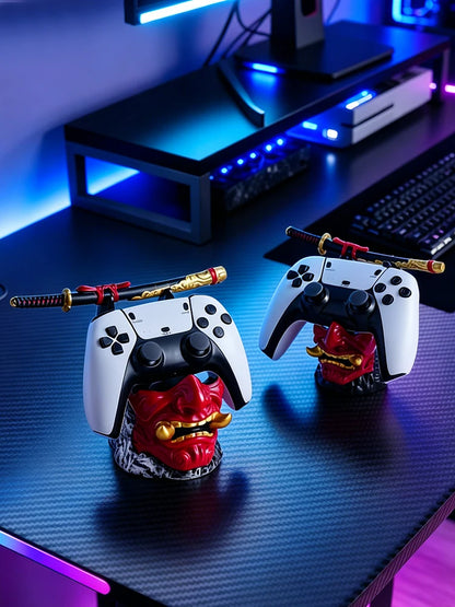 Two red and gold oni mask stands on a desk holding white and black game controllers, each topped with a small samurai sword.
