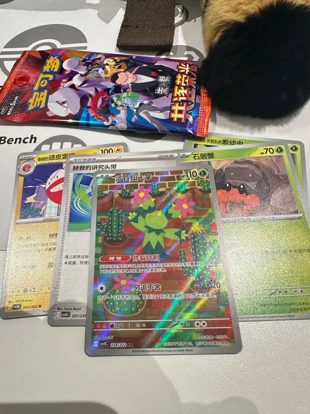 A colorful Pokemon foil card featuring Maractus surrounded by potted cacti, next to other trading cards and a booster pack.