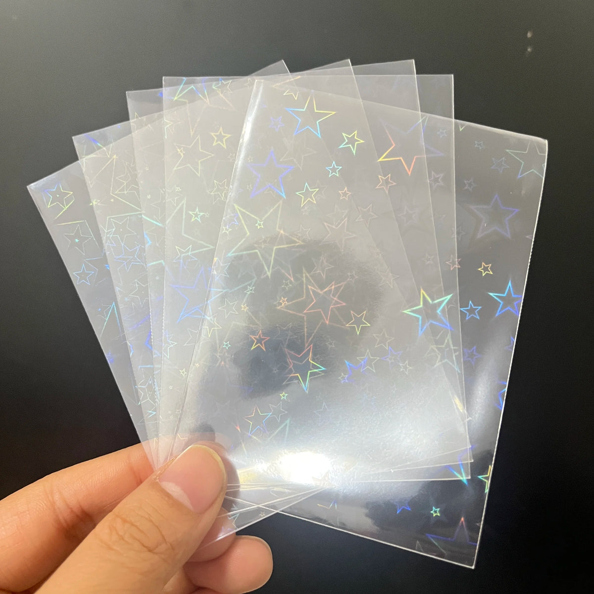 Fanned clear trading card sleeves feature a holographic laser foil star pattern in varying sizes.