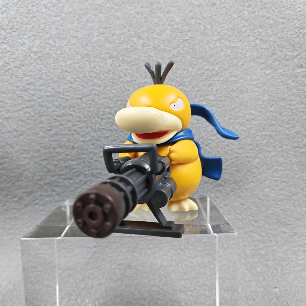Yellow duck-like figurine with a determined expression, wearing a blue scarf and holding a large black Gatling gun.