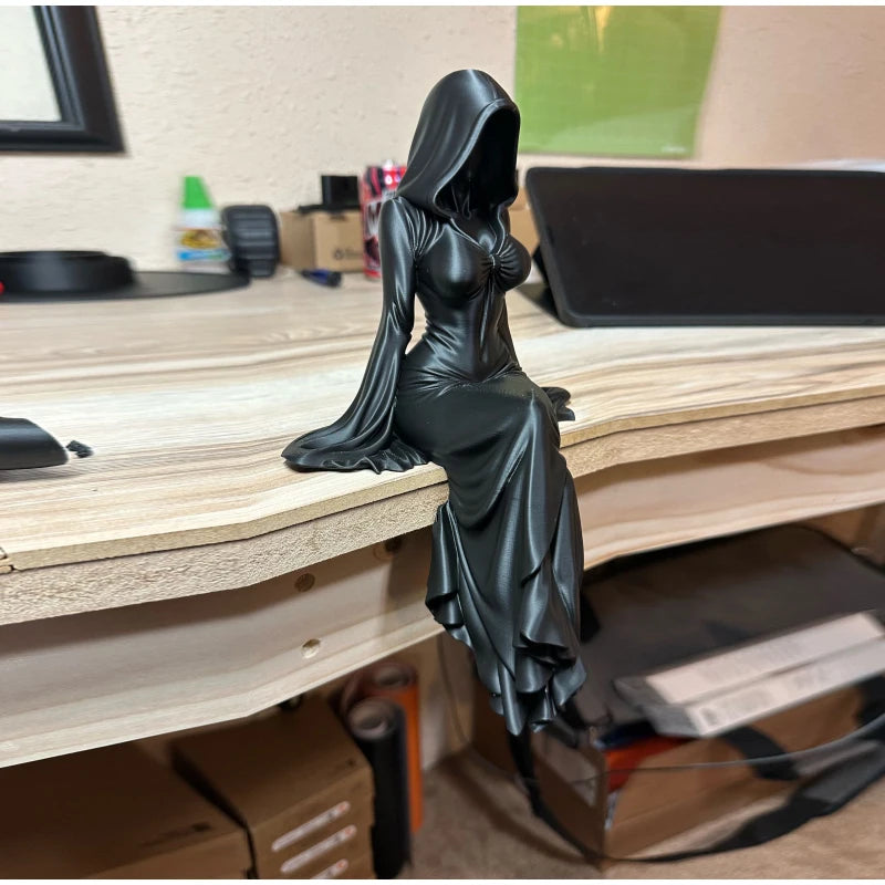 A black, hooded female figurine sits on a wooden desk edge, her long dress draping downward.