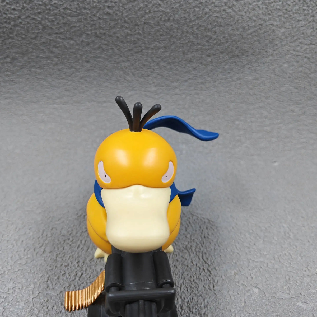 Yellow duck-like figurine with a blue scarf, holding a black Gatling gun, viewed from above.