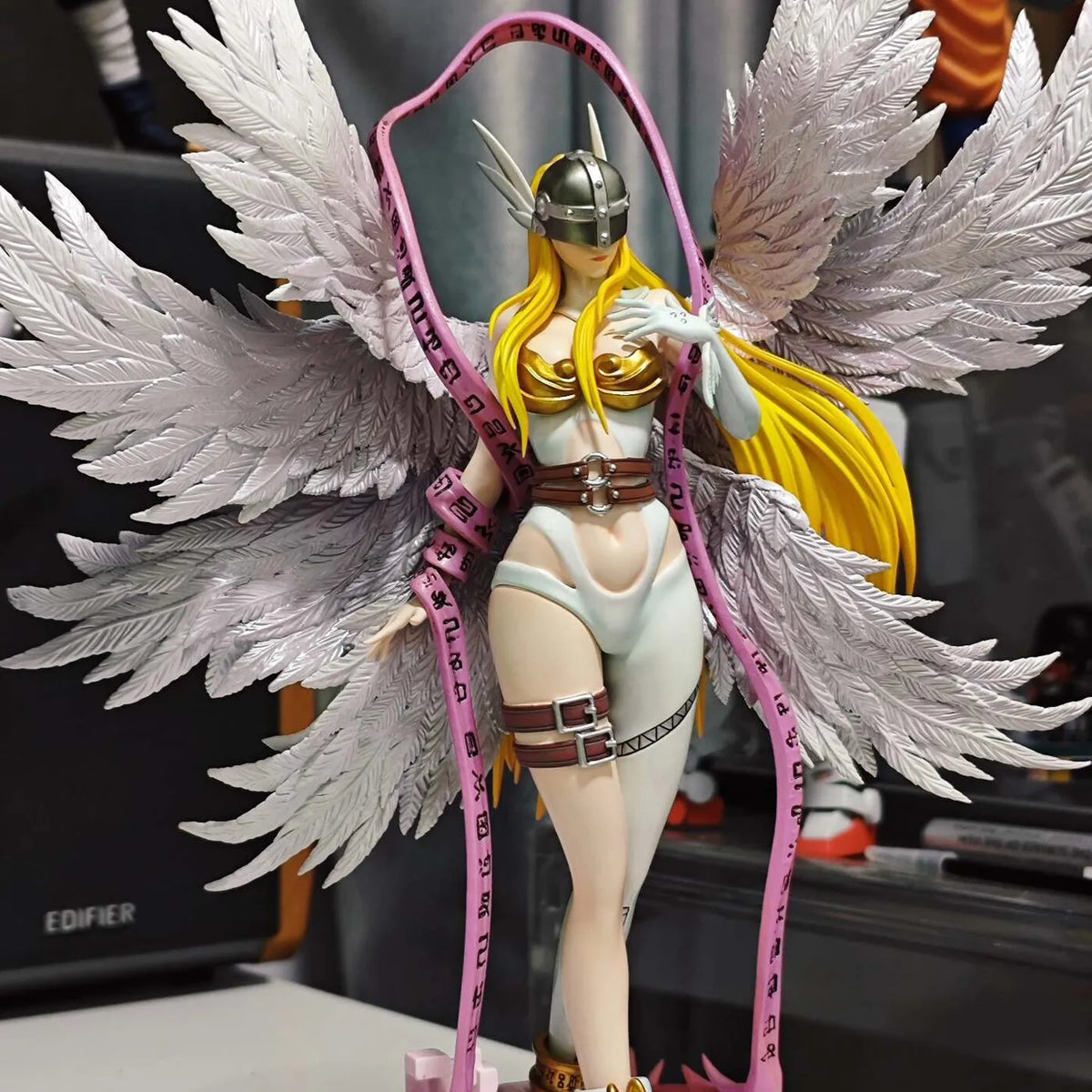 Angewomon PVC statue with large white feathered wings, blonde hair, silver helmet, and a pink rune ribbon.