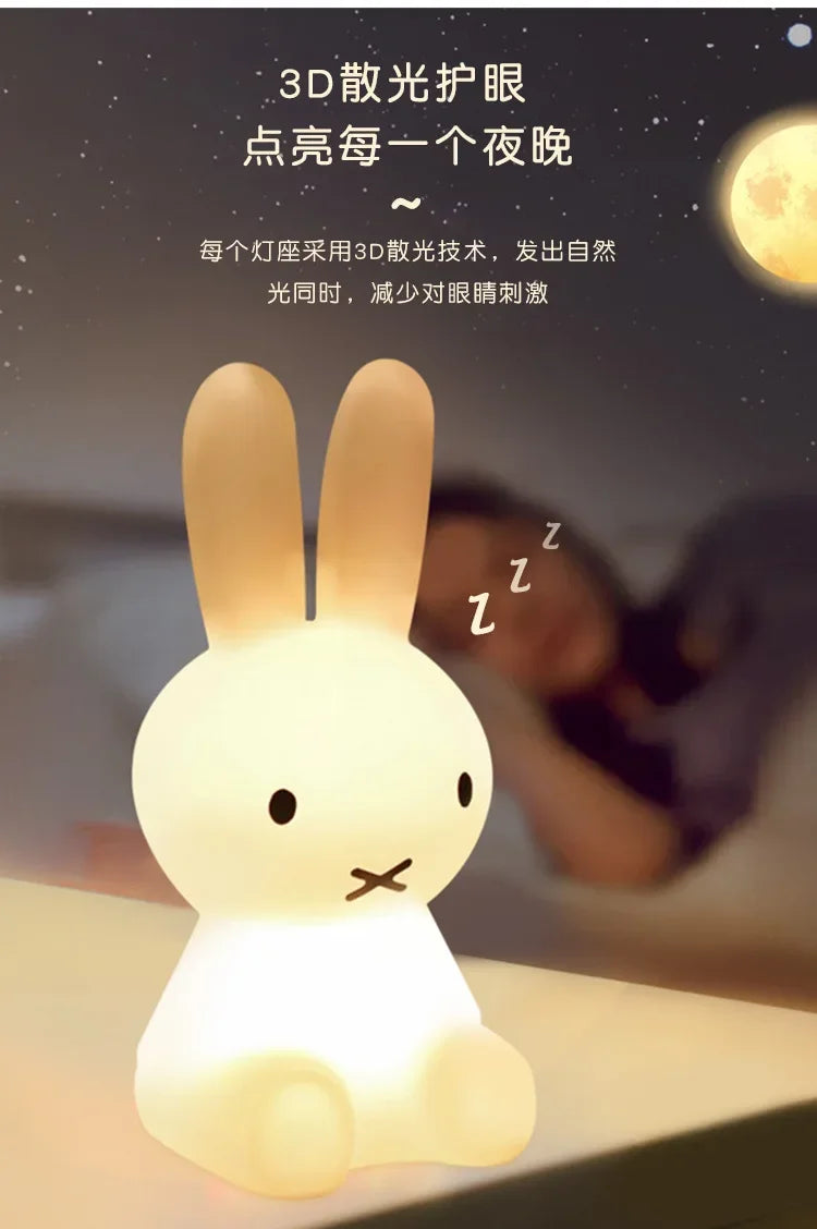 A glowing white Miffy rabbit night light sits on a surface, with a sleeping person blurred in the background.