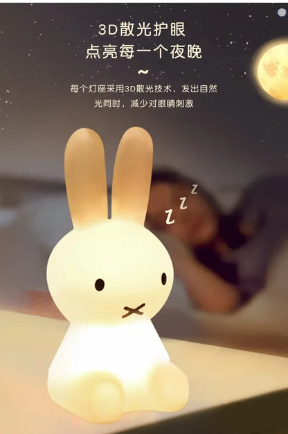 A glowing white Miffy rabbit night light sits on a surface, with a sleeping person blurred in the background.
