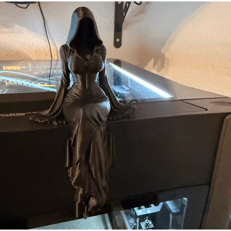 A black hooded female figure with long flowing sleeves sits on a dark surface, legs crossed.