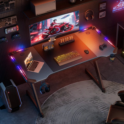 Black gaming desk with carbon-fiber wings, glowing RGB lights, a laptop, monitor, and keyboard.