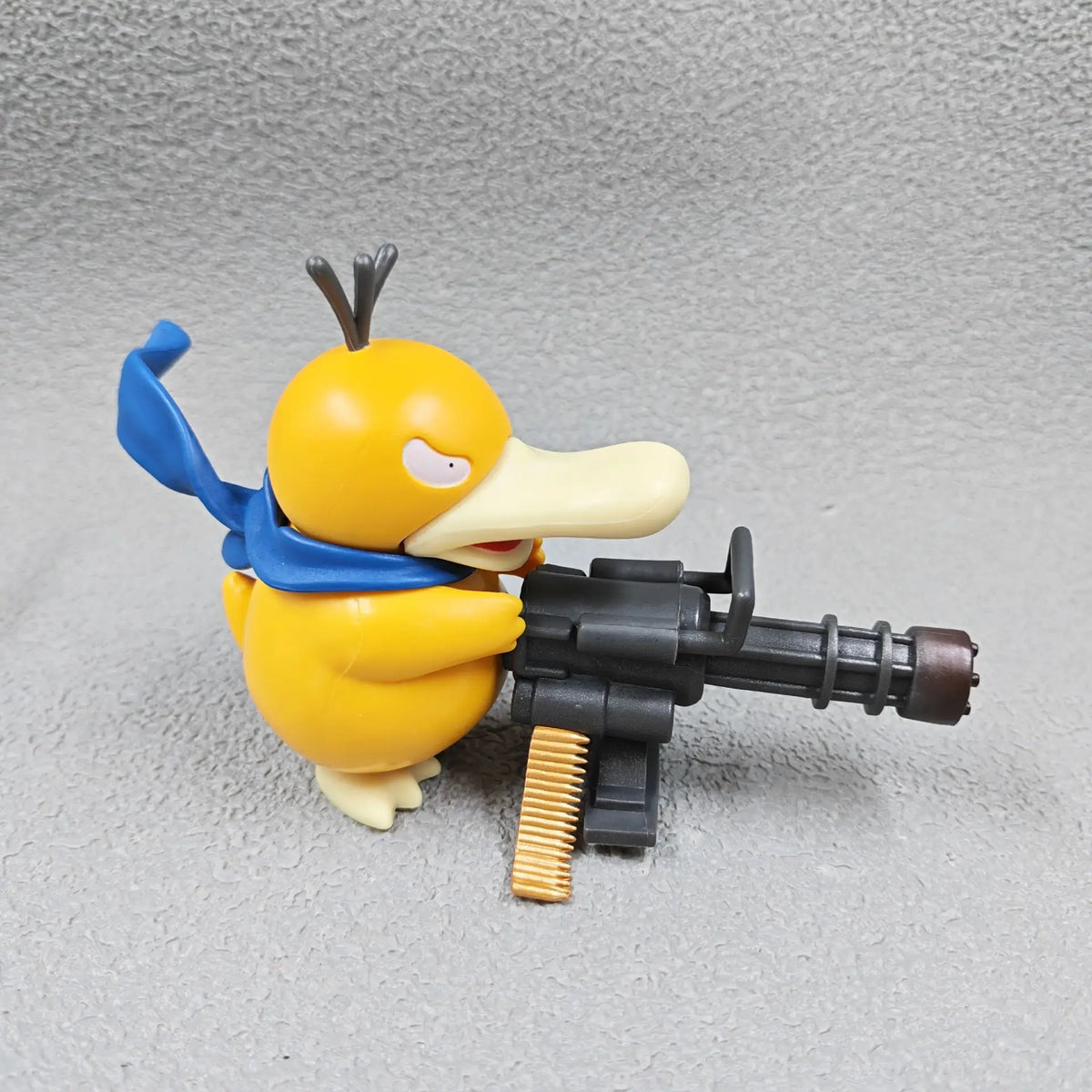 Yellow Psyduck figurine holding a black Gatling gun with a blue scarf on a grey surface.