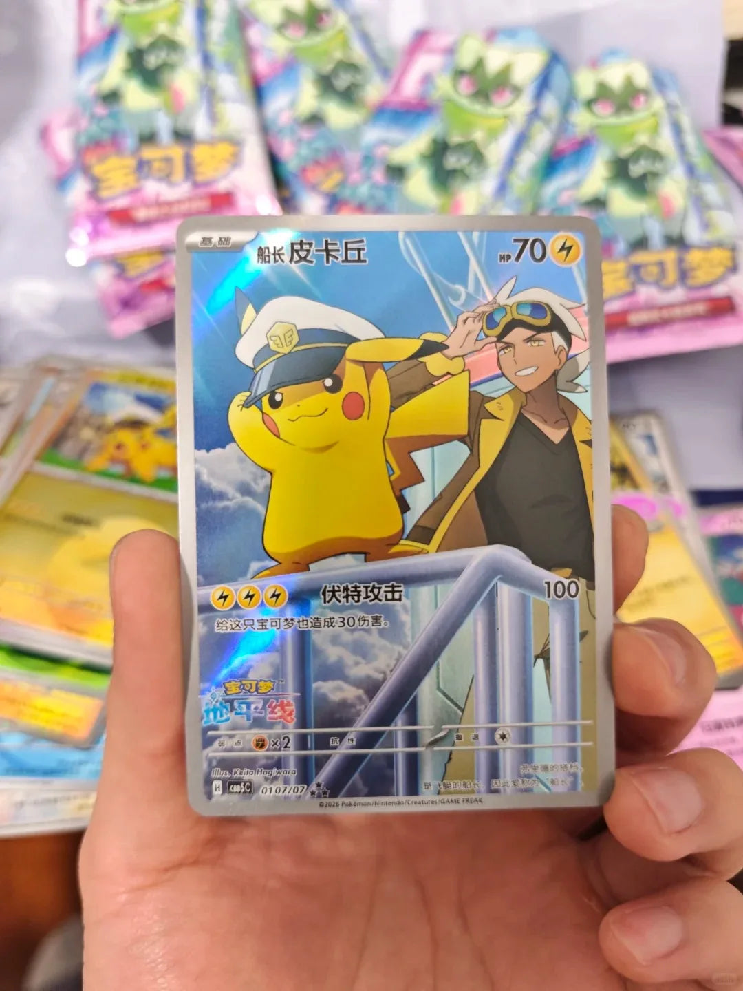 A hand holds a shiny Pokémon card featuring Captain Pikachu in a sailor hat and a trainer against a blue sky.