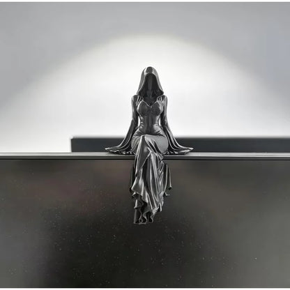 A black figurine of a hooded woman in a fitted dress sitting and dangling her legs.