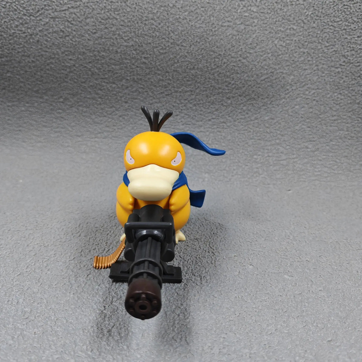 Yellow duck-like figurine with a blue scarf, holding a large black gatling gun, on a grey background.