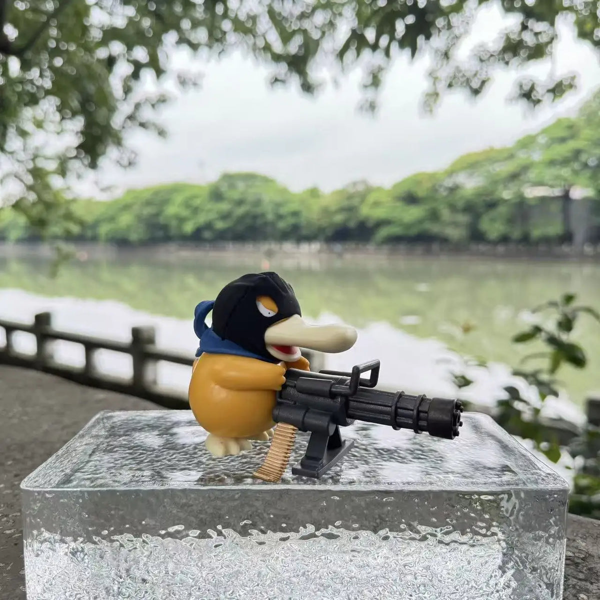 Yellow duck-like figurine with a black ski mask holds a black Gatling gun on a glass block.