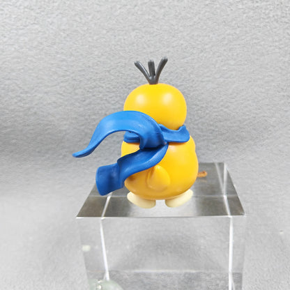 Yellow figurine with a blue scarf and dark hair strands, seen from behind on a clear block.