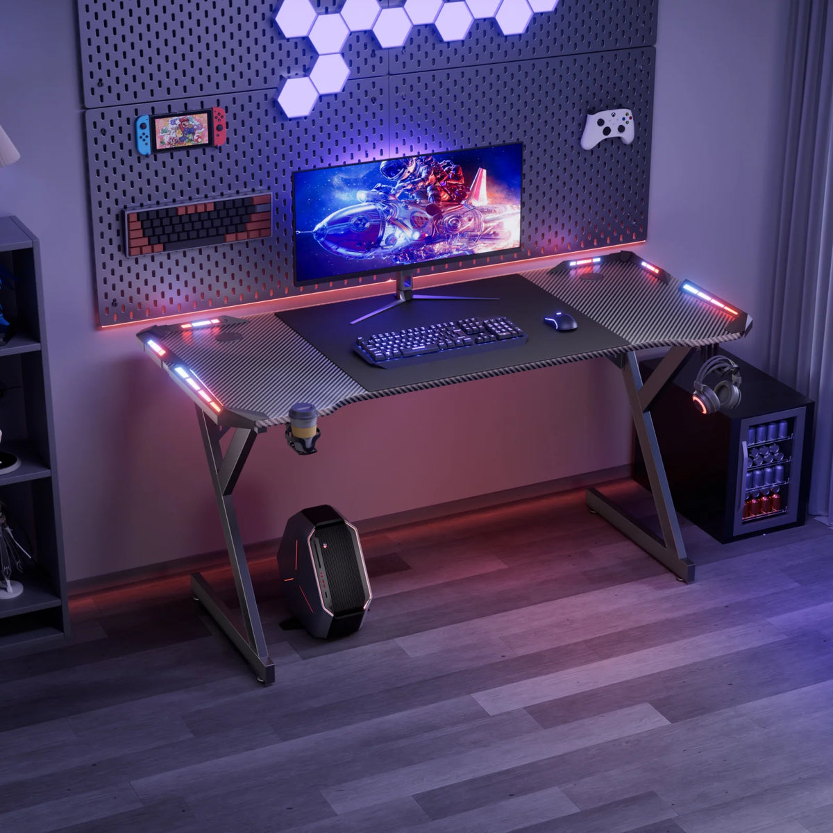 Black carbon-fiber gaming desk with Y-shaped legs, red-and-blue glowing RGB strips, cup holder, and headphone hook.