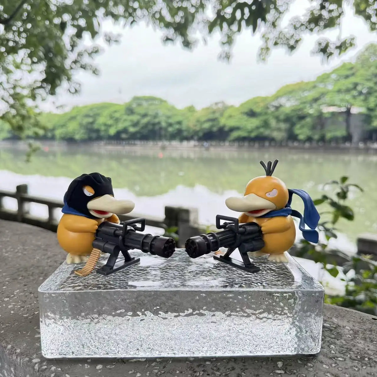 Two yellow Psyduck figurines wearing blue scarves and holding black Gatling guns on a clear block.