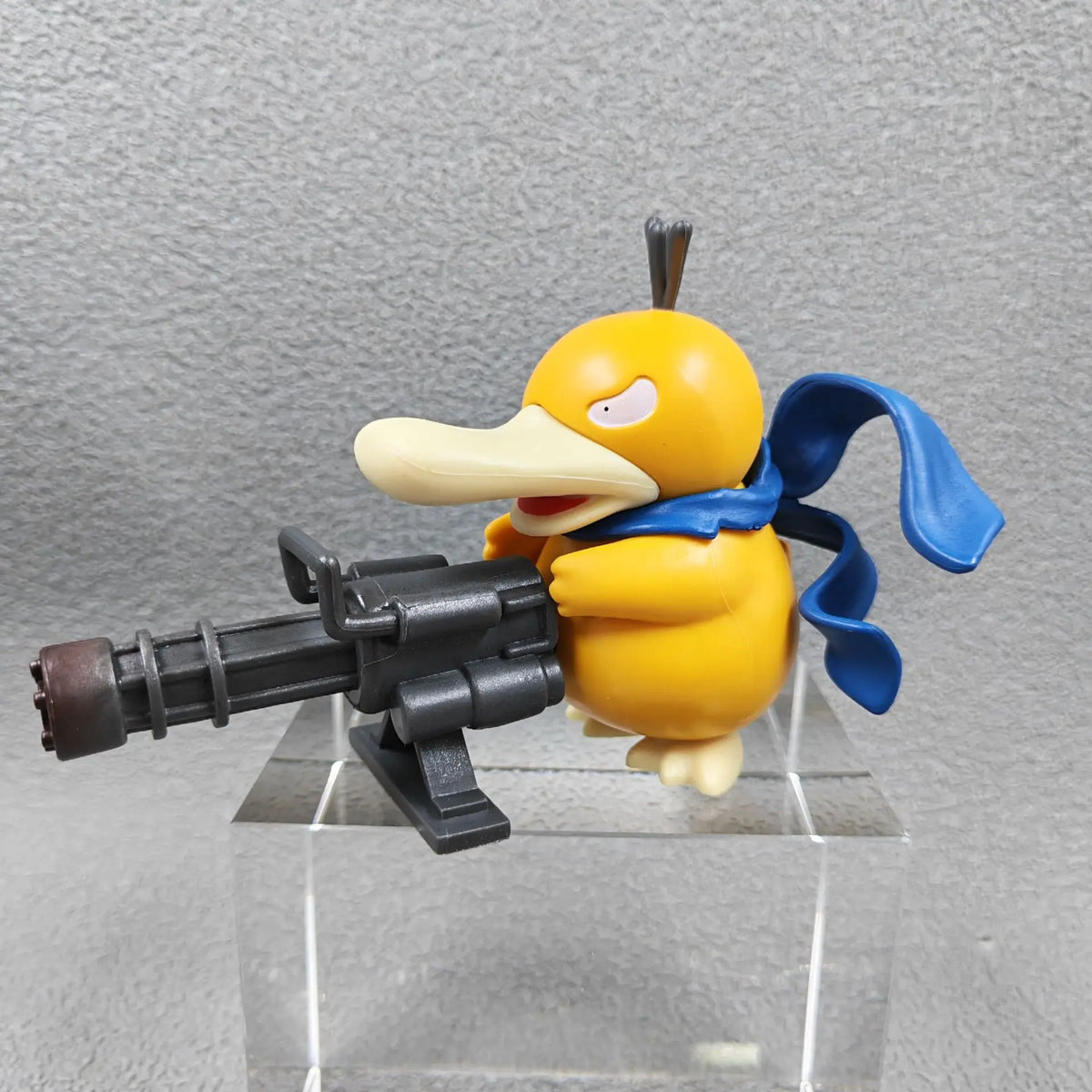 A yellow duck-like figurine with a blue scarf wielding a large black gatling gun.