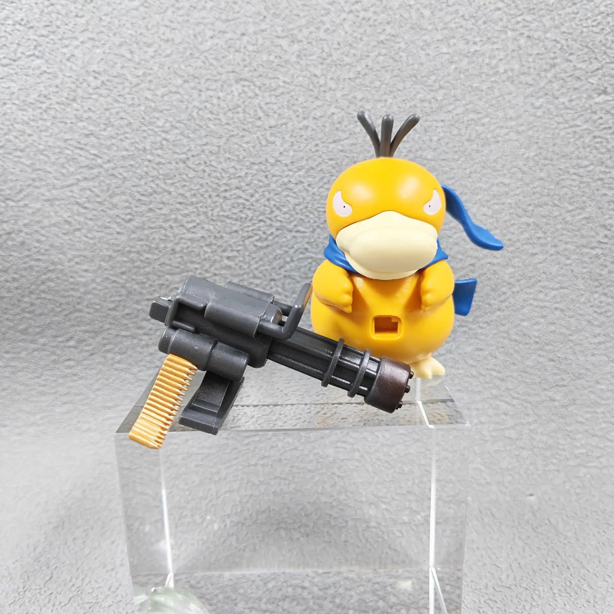 Yellow duck-like figure with a blue scarf standing beside a gray miniature Gatling gun on a clear pedestal.